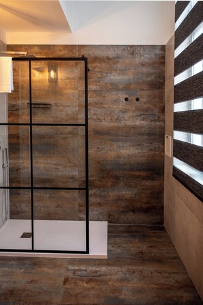 A modern bathroom with a glass shower enclosure, a wooden floor, and a large window providing natural light.