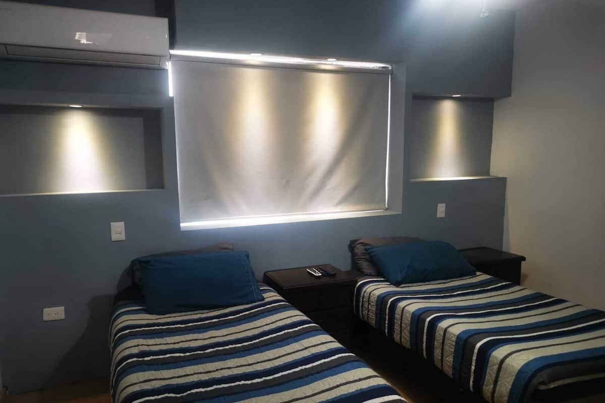 A bedroom with two single beds, a window with blinds, and a ceiling-mounted air conditioning unit.