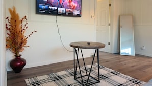 Una sala de estar con un televisor de pantalla plana montado en la pared, una mesa redonda de madera, un tapete estampado y un jarrón con plantas secas.