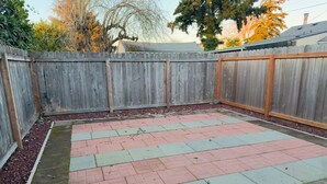 Un patio trasero con cerca de madera, un área de patio empedrada con baldosas rojas y verdes, y un macizo de jardín con acolchado rojo.