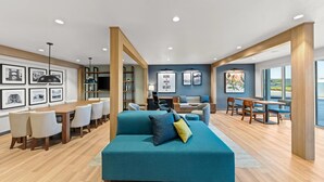 A spacious living area with a large teal sofa, wooden dining table, and chairs. There are framed pictures on the walls and a large window providing a view of the outdoors. - Everhome Suites Waco by Choice Hotels (Waco)