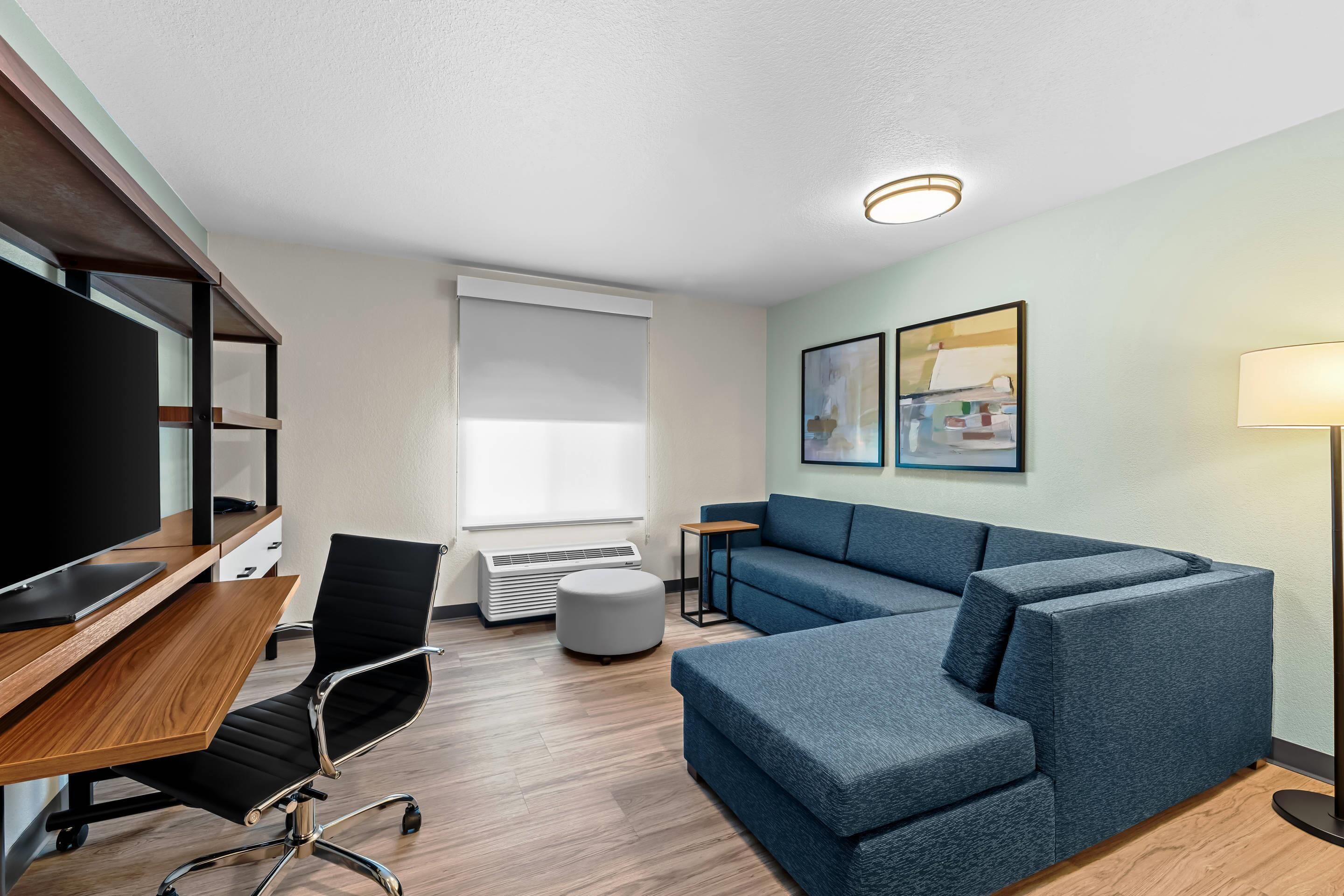 Une chambre d’hôtel moderne avec un bureau en bois, un fauteuil de bureau noir, un canapé bleu et un téléviseur à écran plat.