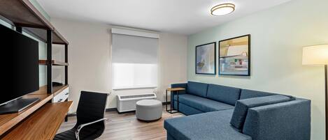Une chambre d’hôtel moderne avec un bureau en bois, un fauteuil de bureau noir, un canapé bleu et un téléviseur à écran plat.