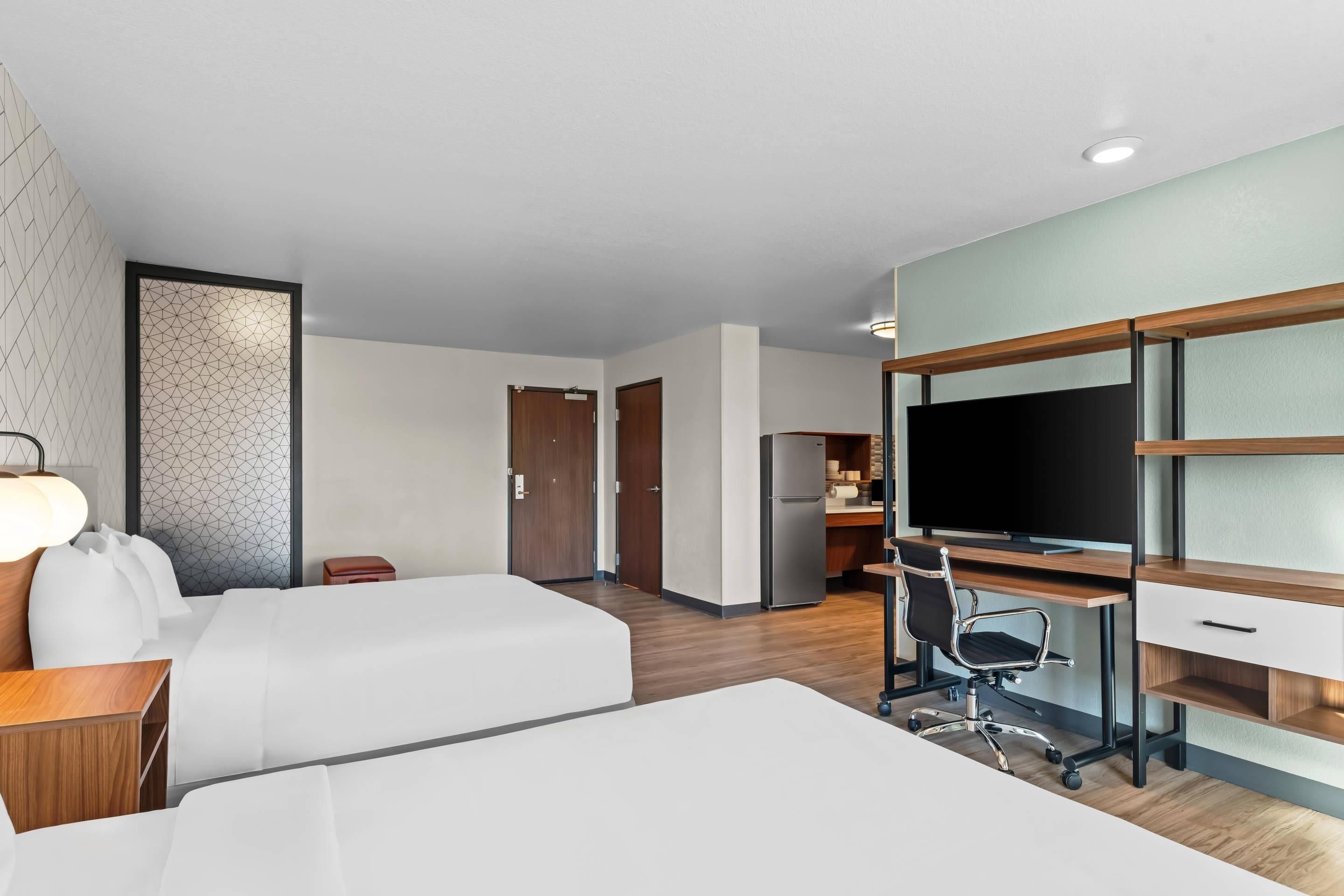 Ein Hotelzimmer mit zwei Betten, einem Schreibtisch mit Computer, einem Fernseher und einer Küchenzeile.