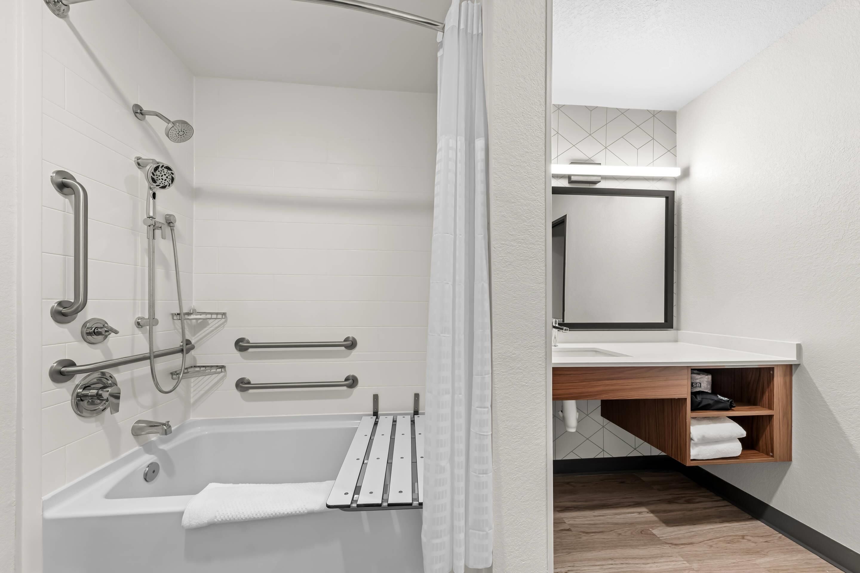 A modern bathroom with a walk-in shower, grab bars, a bathtub, and a wooden vanity with a mirror.
