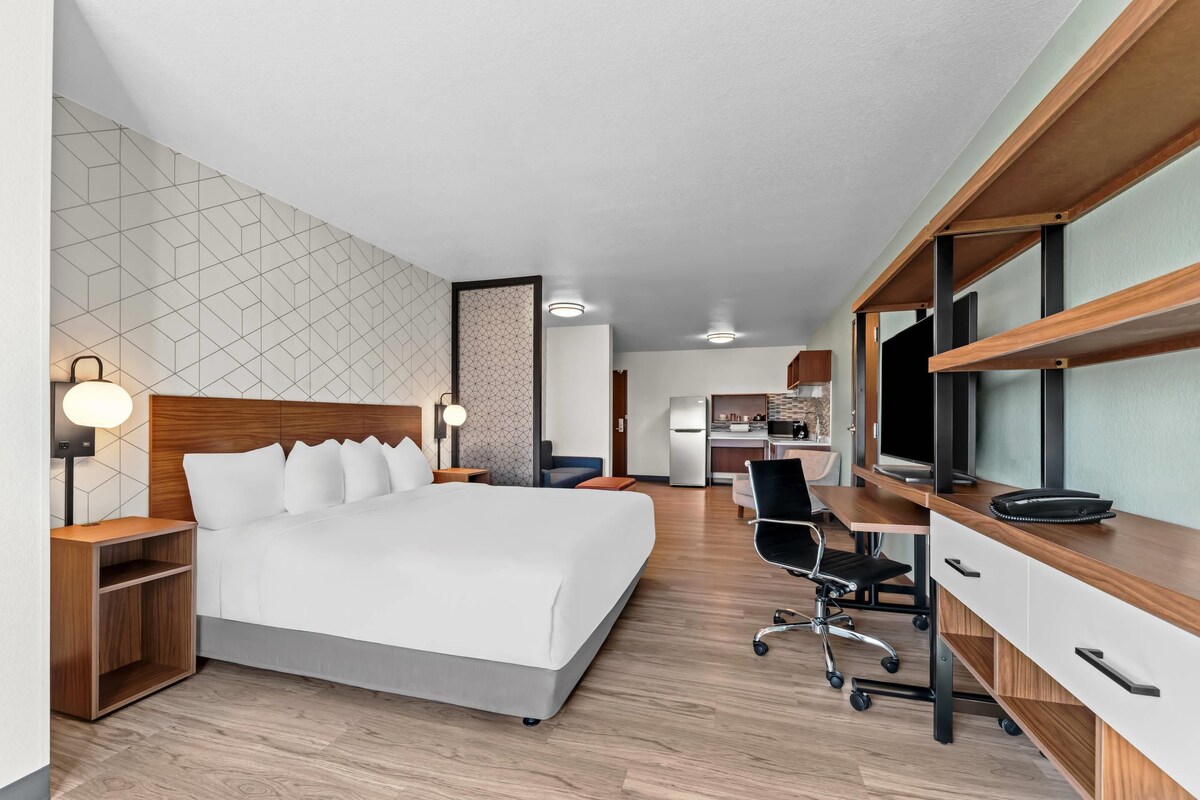 A modern hotel room with a neatly made bed, a desk with a computer, and a kitchenette in the background.