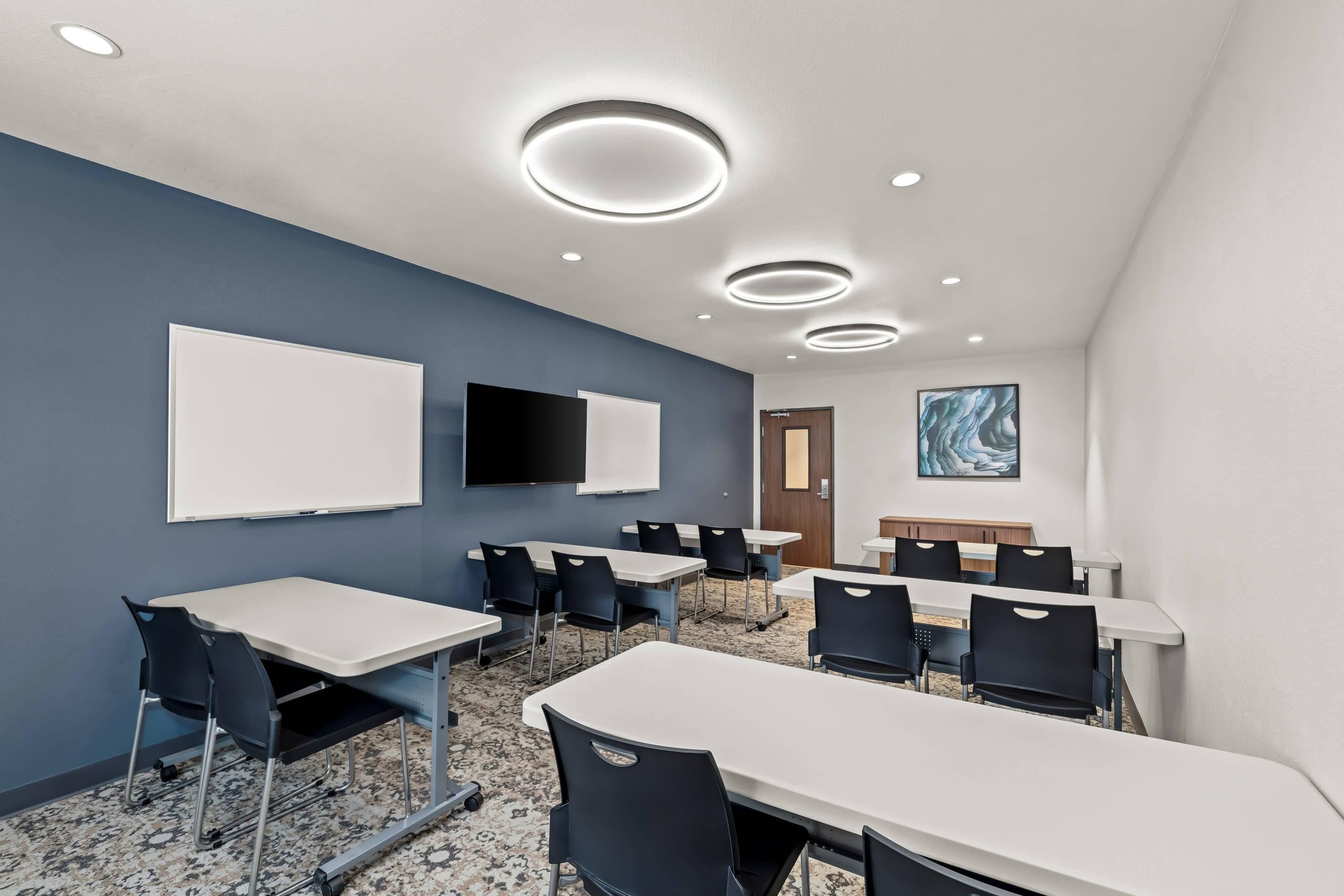 A conference room with white tables and chairs, a whiteboard, a flat-screen TV, and a painting on the wall.
