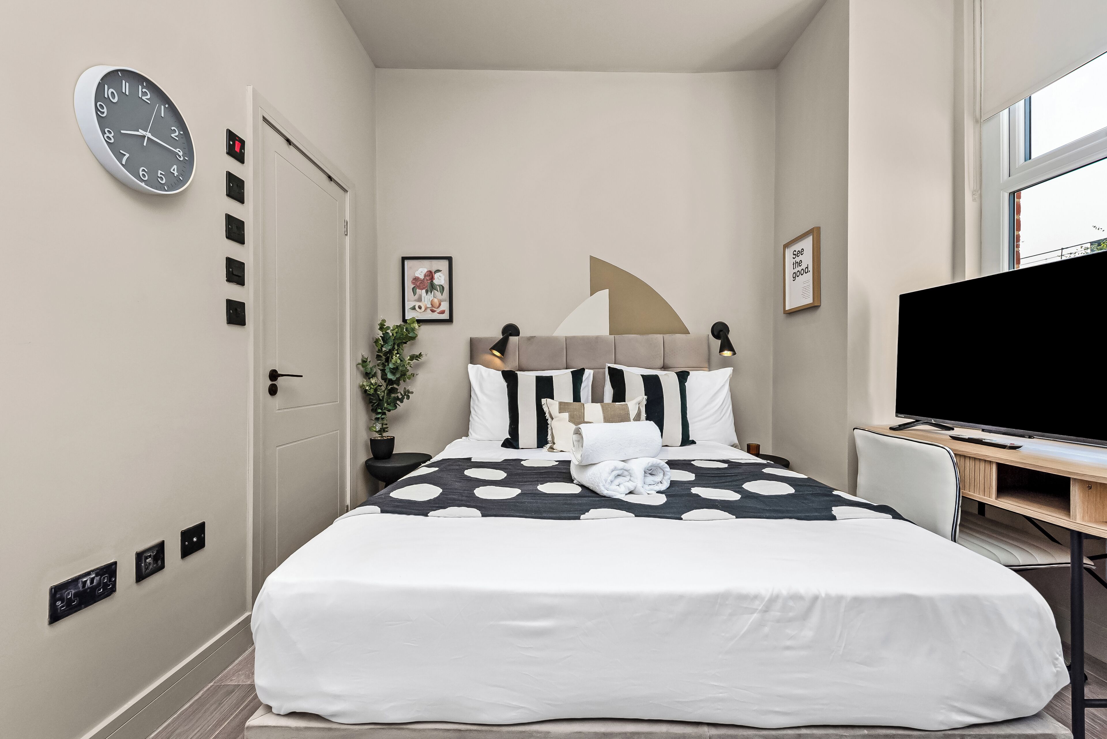 Una camera d'albergo moderna con un letto, una TV a schermo piatto, una sveglia e una finestra.