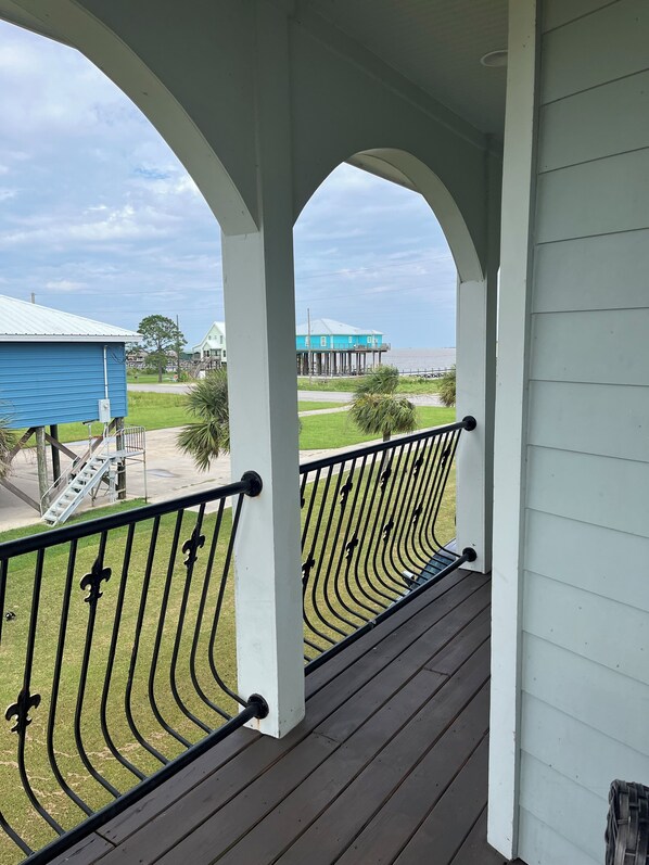 A balcony with a metal railing, white pillars, and a view of a blue building and a pier in the distance.
