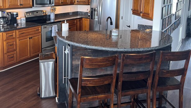 A spacious kitchen with wooden cabinets, a stainless steel refrigerator, a granite island with bar stools, and pendant lighting. - LAKE HOUSE. Serene, charming 5-bedroom house in greater New Orleans. (New Orleans)