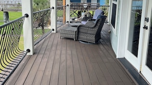A covered porch with a wooden deck, wicker furniture, and a view of a blue house in the background. - LAKE HOUSE. Serene, charming 5-bedroom house in greater New Orleans. (New Orleans)