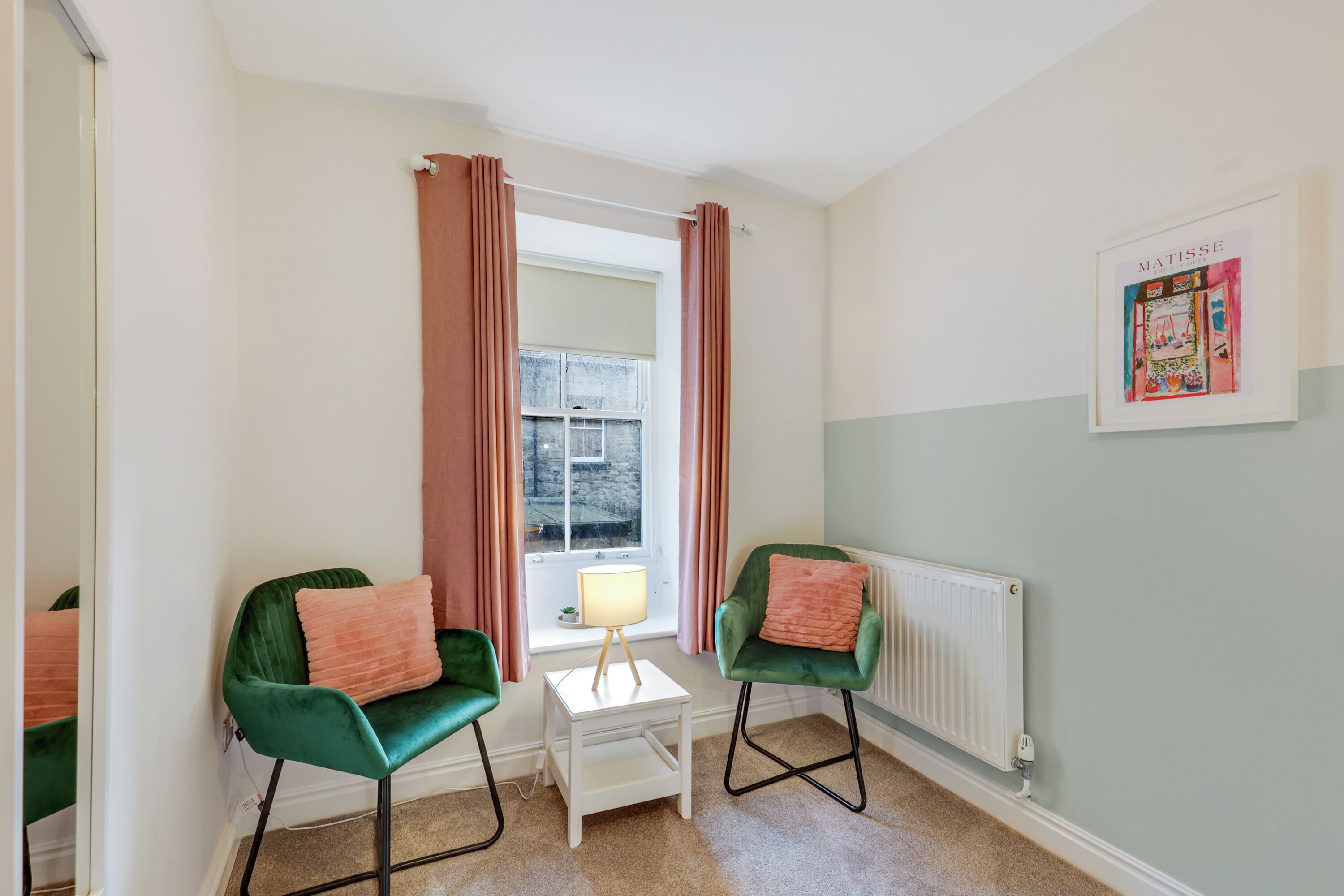 Une pièce avec deux chaises vertes, une petite table blanche avec une lampe, un radiateur et une fenêtre avec des rideaux.