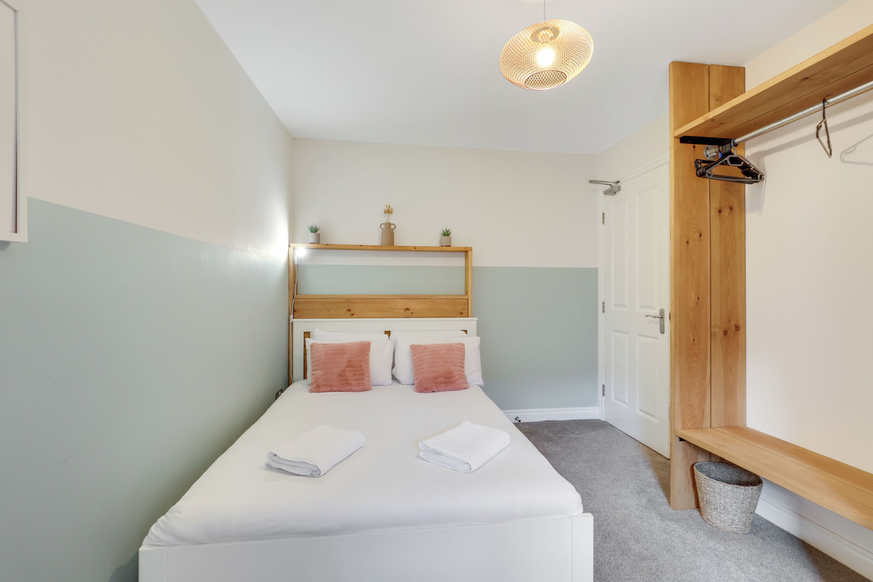 Una cama bien tendida con dos almohadas y dos toallas dobladas, un cabecero de madera, una pequeña estantería con objetos decorativos, una lámpara colgante y un banco de madera con una cesta debajo.