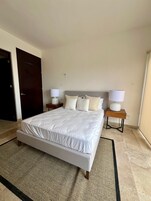 Una cama bien hecha con sábanas blancas, dos mesitas de noche con lámparas y una puerta oscura en una habitación de hotel.