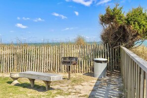 A wooden bench, a trash bin, and a metal sign post in a grassy area with a wooden fence and trees in the background. - Top Floor Beachfront w Pool & Ocean Sunrise Views! (Carolina Beach)