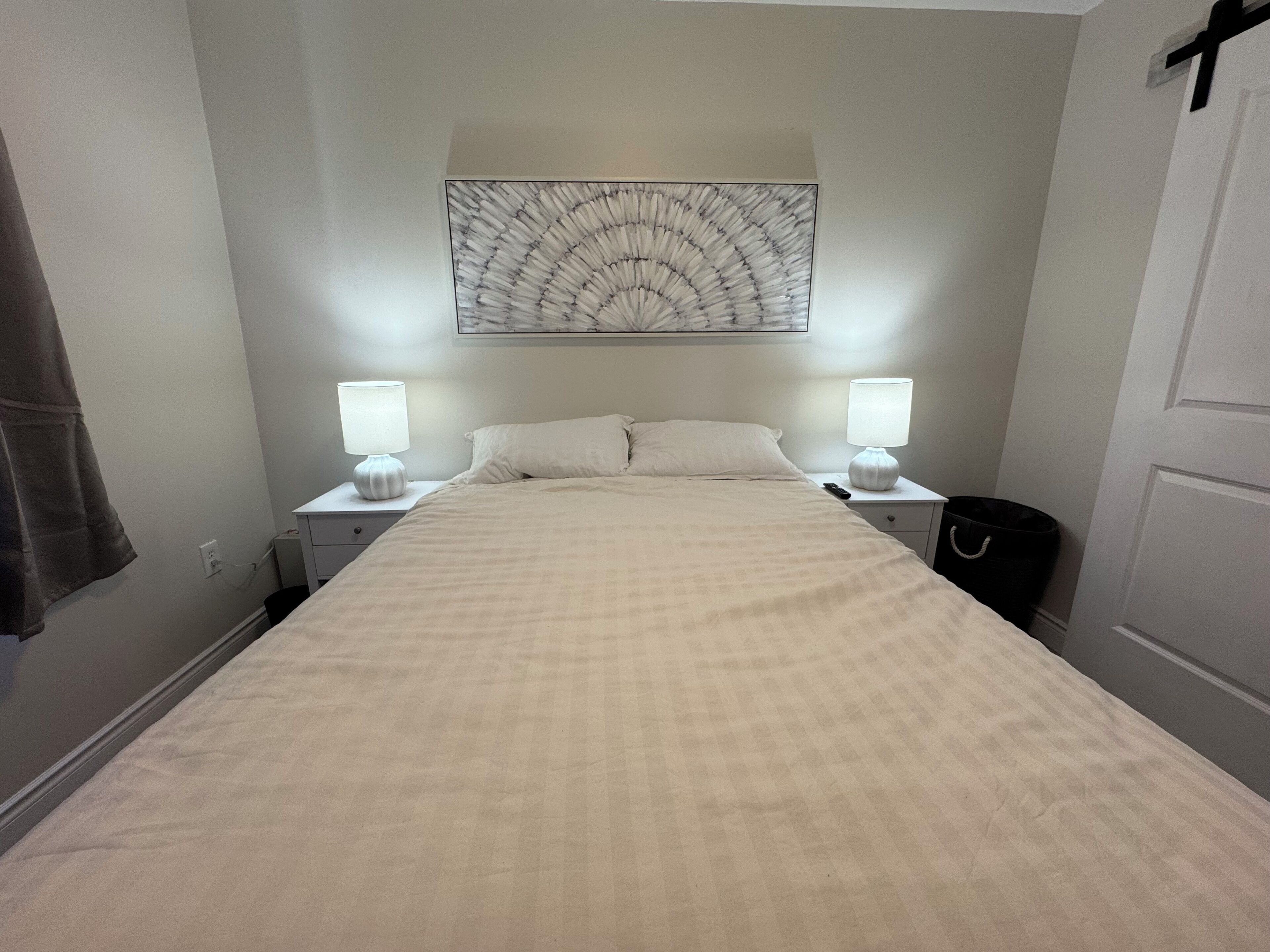 Una cama bien hecha con colcha a cuadros, dos lámparas de noche, una mesita de noche y una obra de arte colgada en la pared sobre la cama.