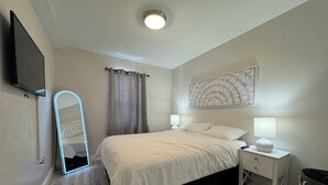 A bedroom with a bed, two bedside tables with lamps, a wall-mounted TV, a large mirror, a ceiling light, and a floor lamp. - Unwind at Windemere 3-Bedroom Apartment in Thunder Bay (Thunder Bay)