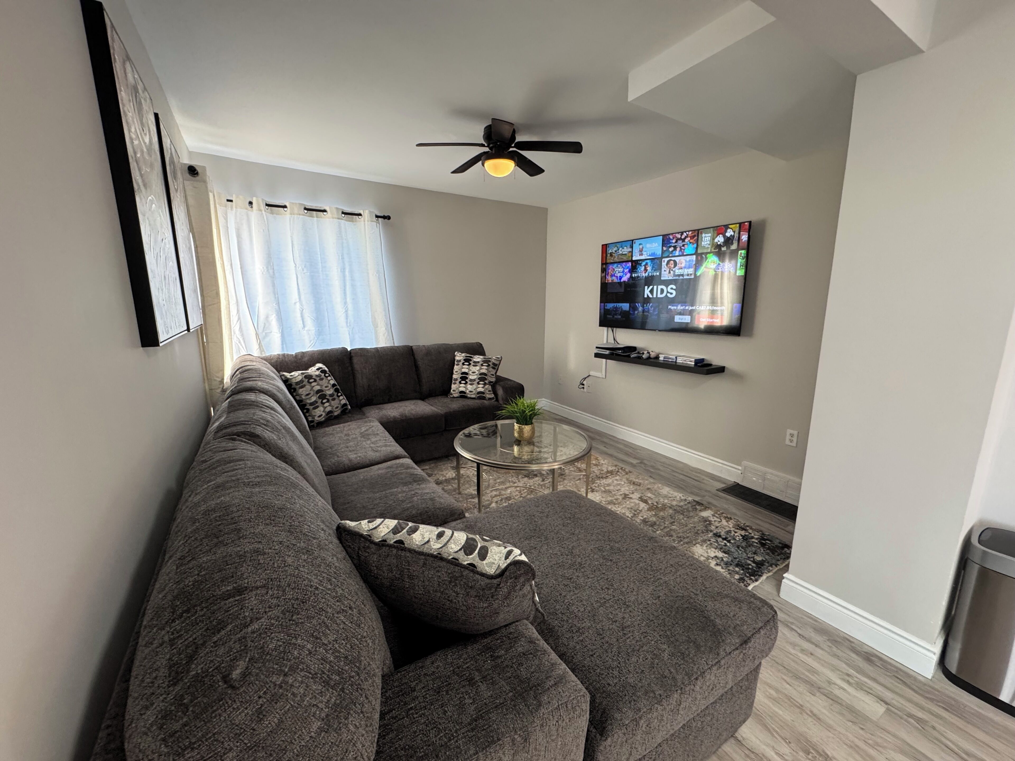 A modern living room with a sectional sofa, a glass coffee table, a ceiling fan, and a flat-screen TV mounted on the wall.