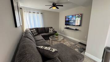 A modern living room with a sectional sofa, a glass coffee table, a ceiling fan, and a flat-screen TV mounted on the wall.