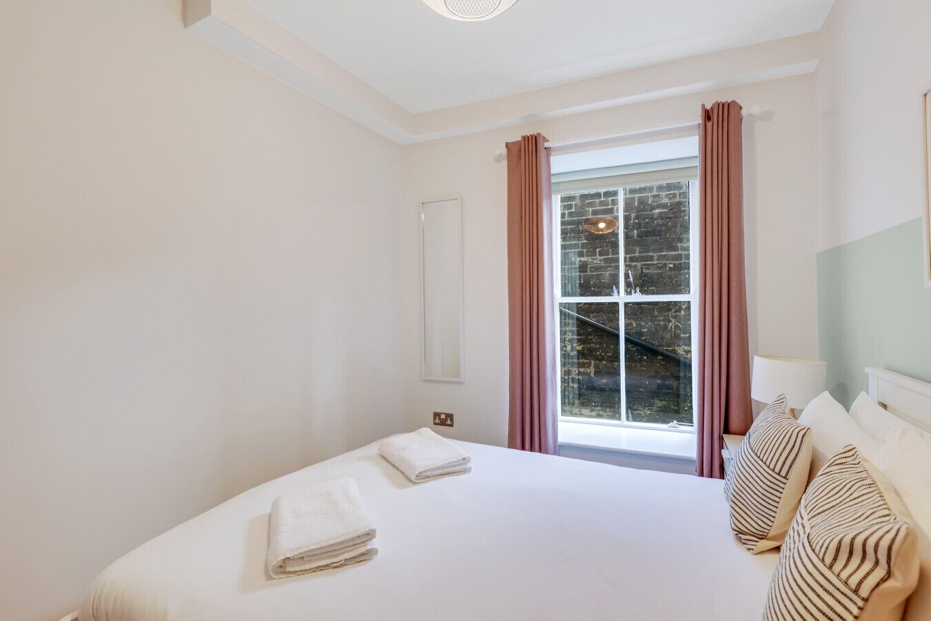 A hotel room with a large bed, a window with curtains, a bedside table with a lamp, and a view of a brick wall outside.