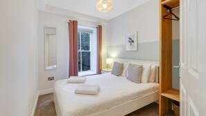 Um quarto com cama, janela com cortinas, abajur e prateleira de madeira.
