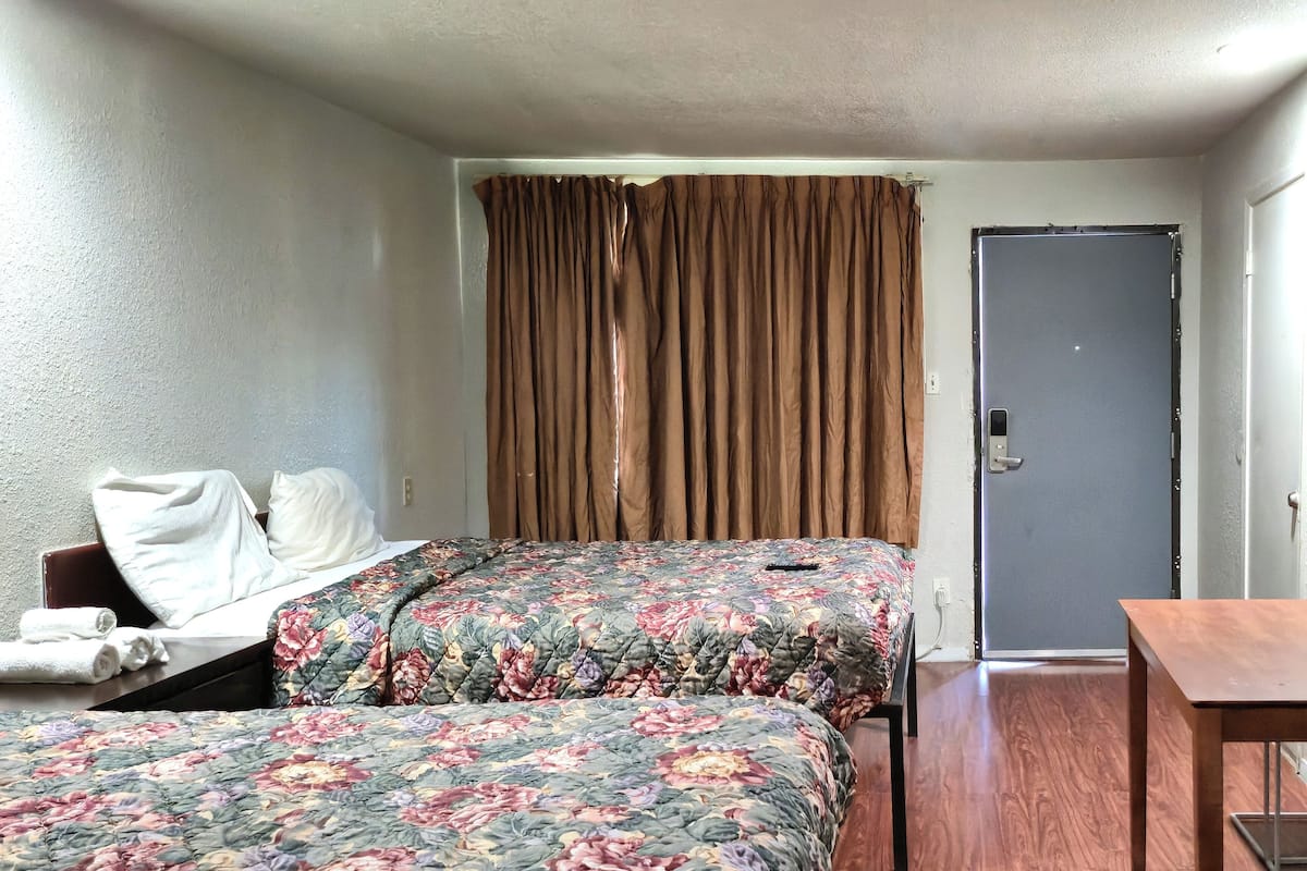 Quarto de hotel com duas camas, uma mesa de madeira, uma porta e uma janela com cortinas.