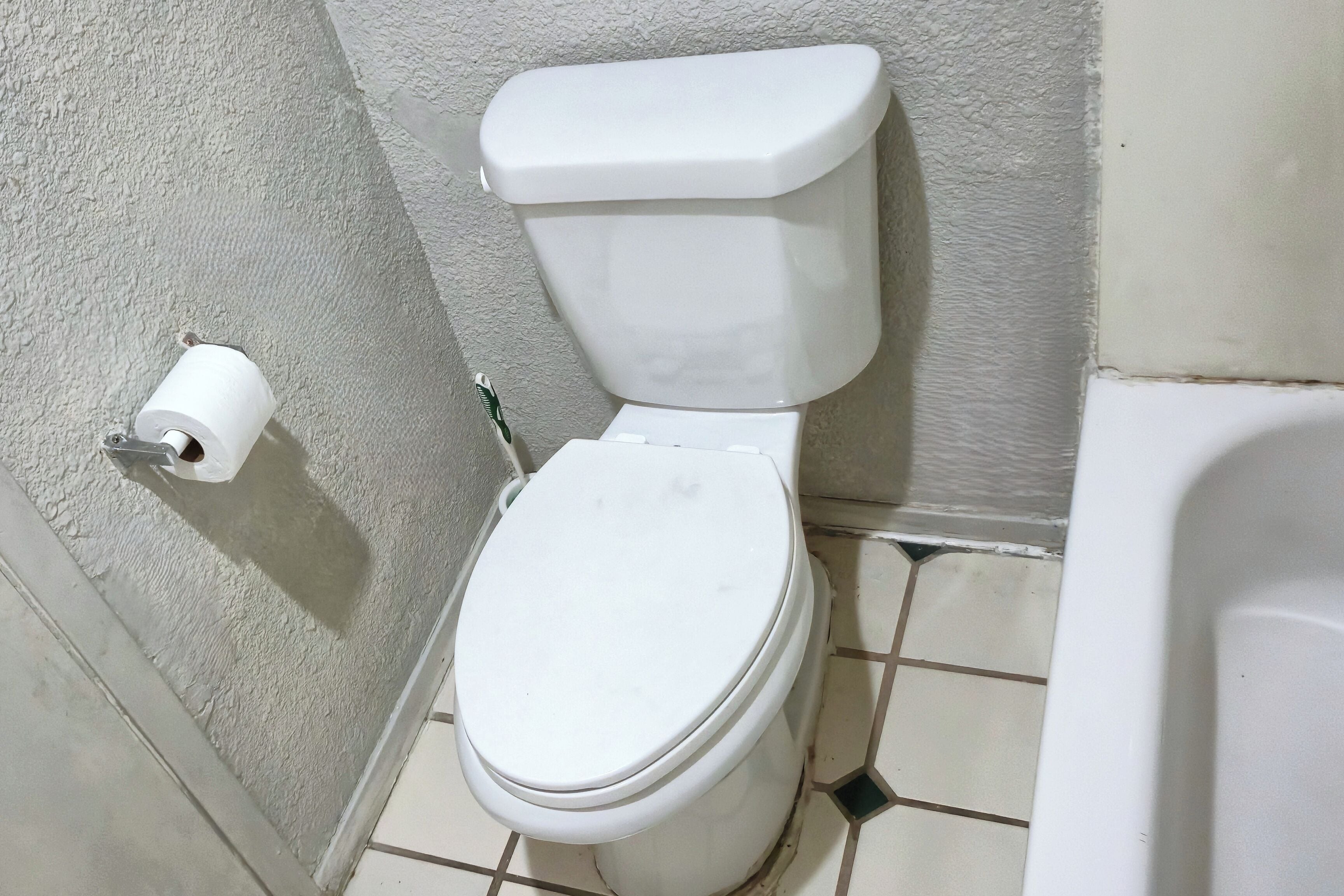 A white toilet with a paper towel holder on the wall, a tiled floor, and a bathtub in the corner.