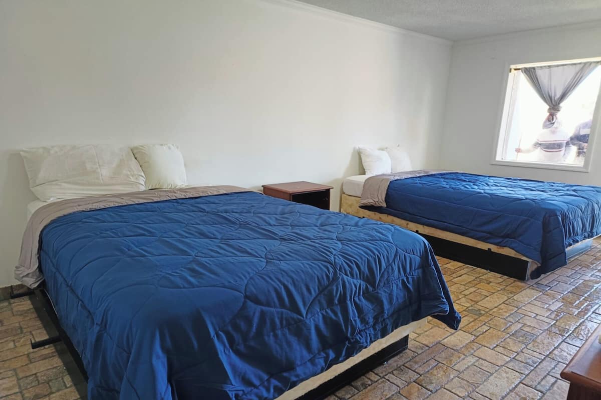 A room with two beds, each with a blue bedspread, a small wooden nightstand, and a window with a view of the outside.