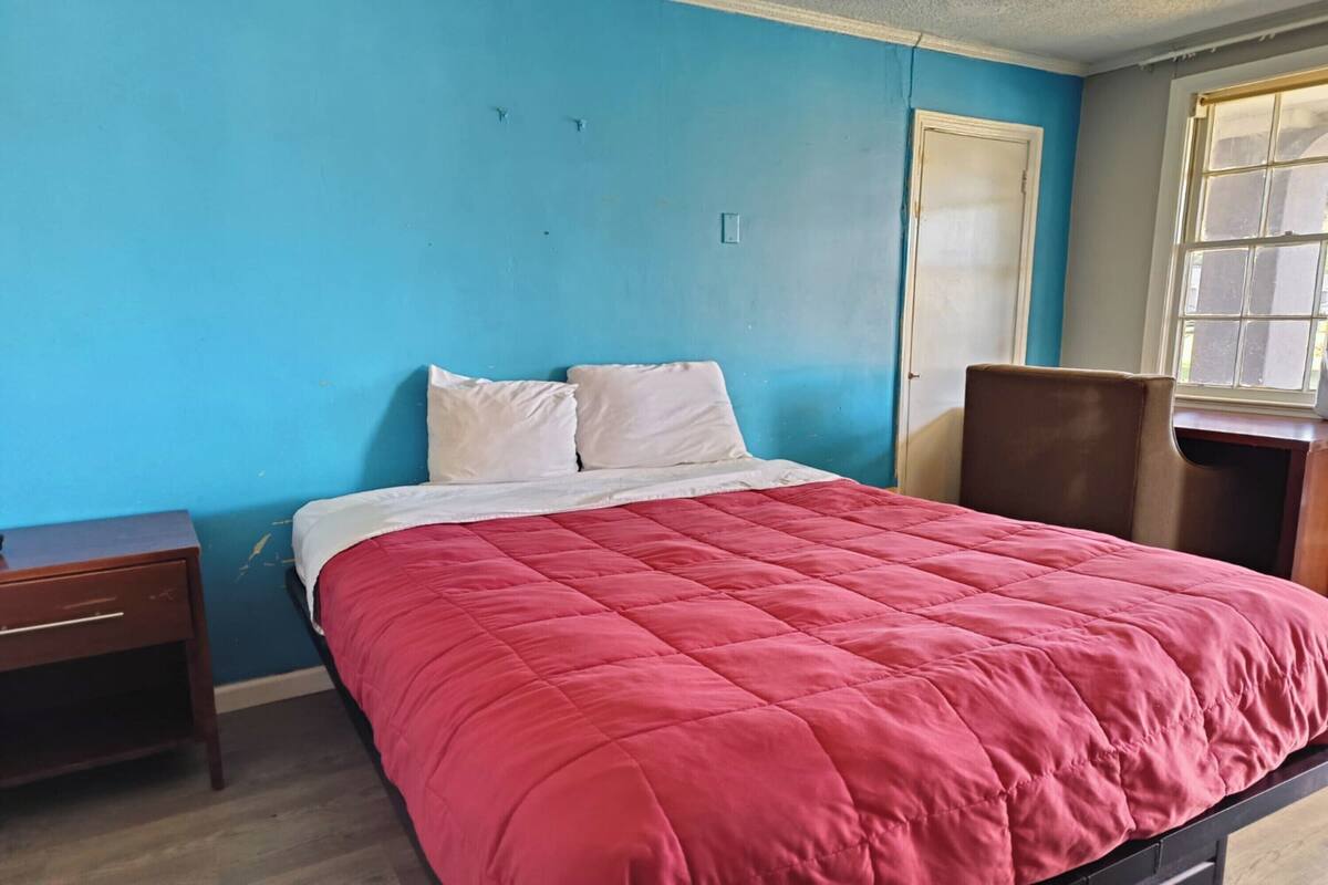 A room with a bed, a wooden nightstand, a chair, and a window.