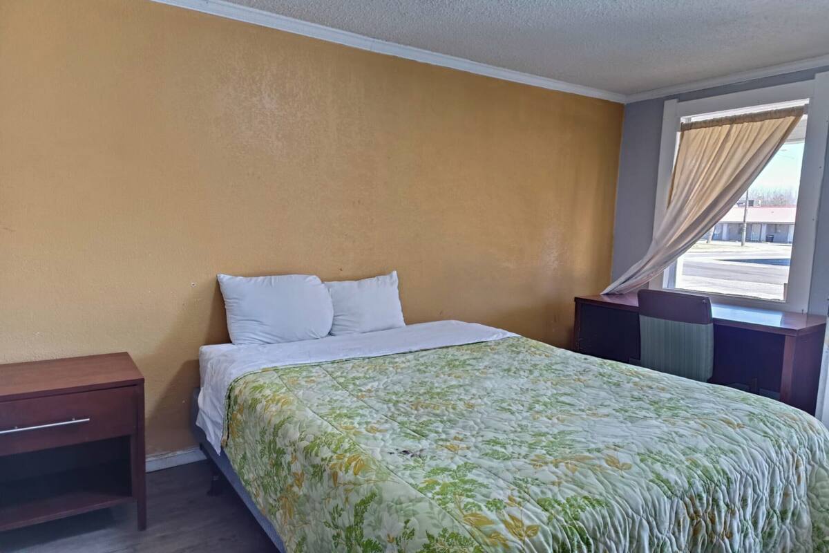 A hotel room with a bed, a nightstand, a window with curtains, and a view of the outside.