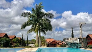 A swimming pool with clear blue water, surrounded by palm trees and buildings with red roofs. - Beautiful and cozy house! (Novo Gravatá Gravatá)