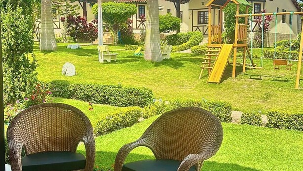 A covered patio area with wicker furniture and a view of a well-maintained garden with a playground. - Beautiful and cozy house! (Novo Gravatá Gravatá)