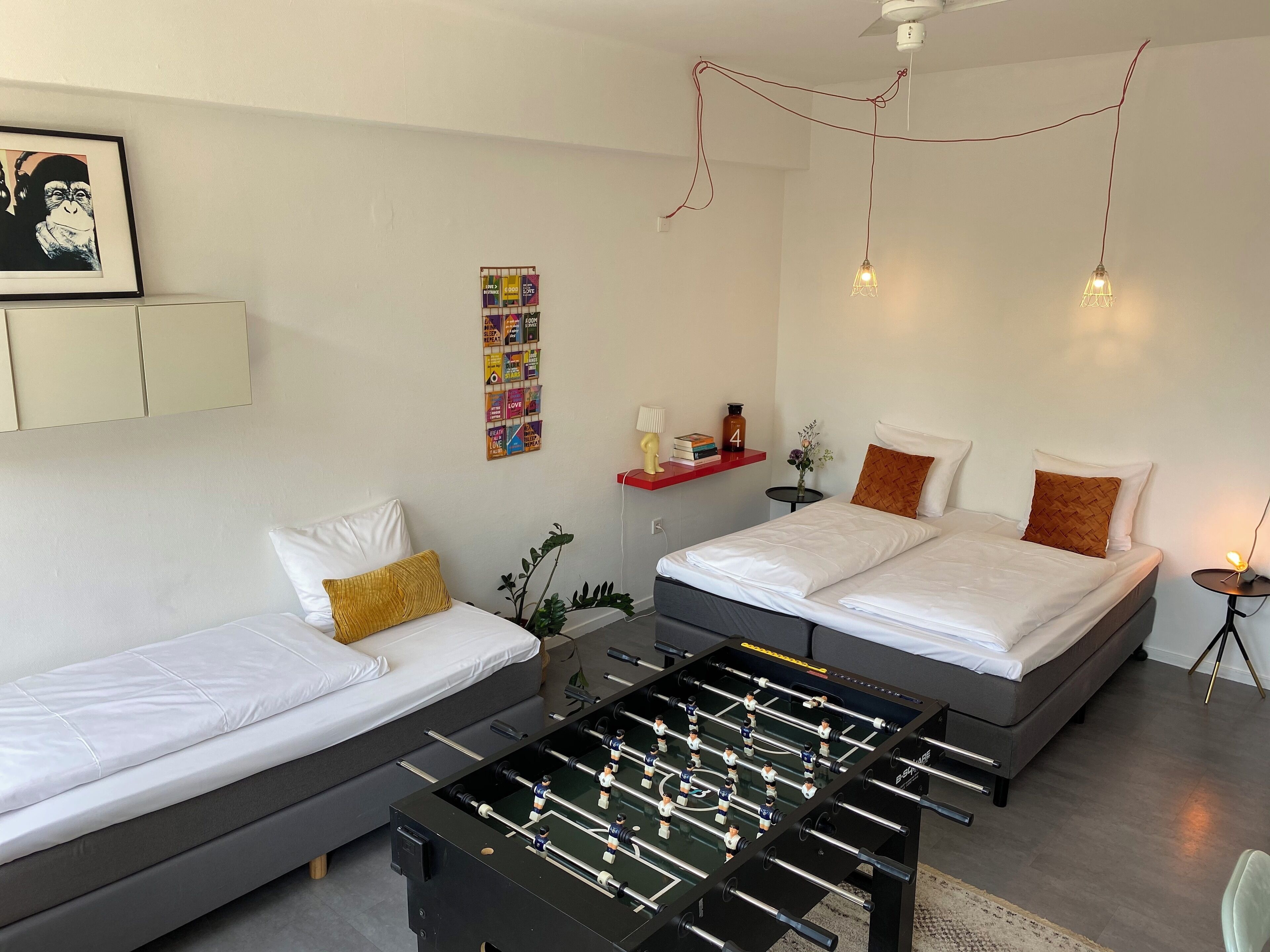 Una habitación de hotel moderna con dos camas, una mesa de futbolín, una mesita con una lámpara y una estantería en la pared con libros y un reloj.