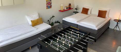 Una habitación de hotel moderna con dos camas, una mesa de futbolín, una mesita con una lámpara y una estantería en la pared con libros y un reloj.