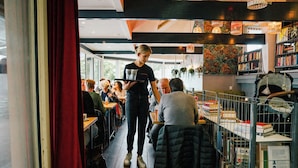 Ein gemütliches Café mit Holztischen, einer Bücherwand und hängenden Pflanzen.
