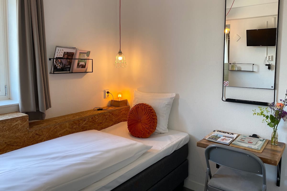 A hotel room with a bed, bedside table, lamp, mirror, and a small desk with a chair.