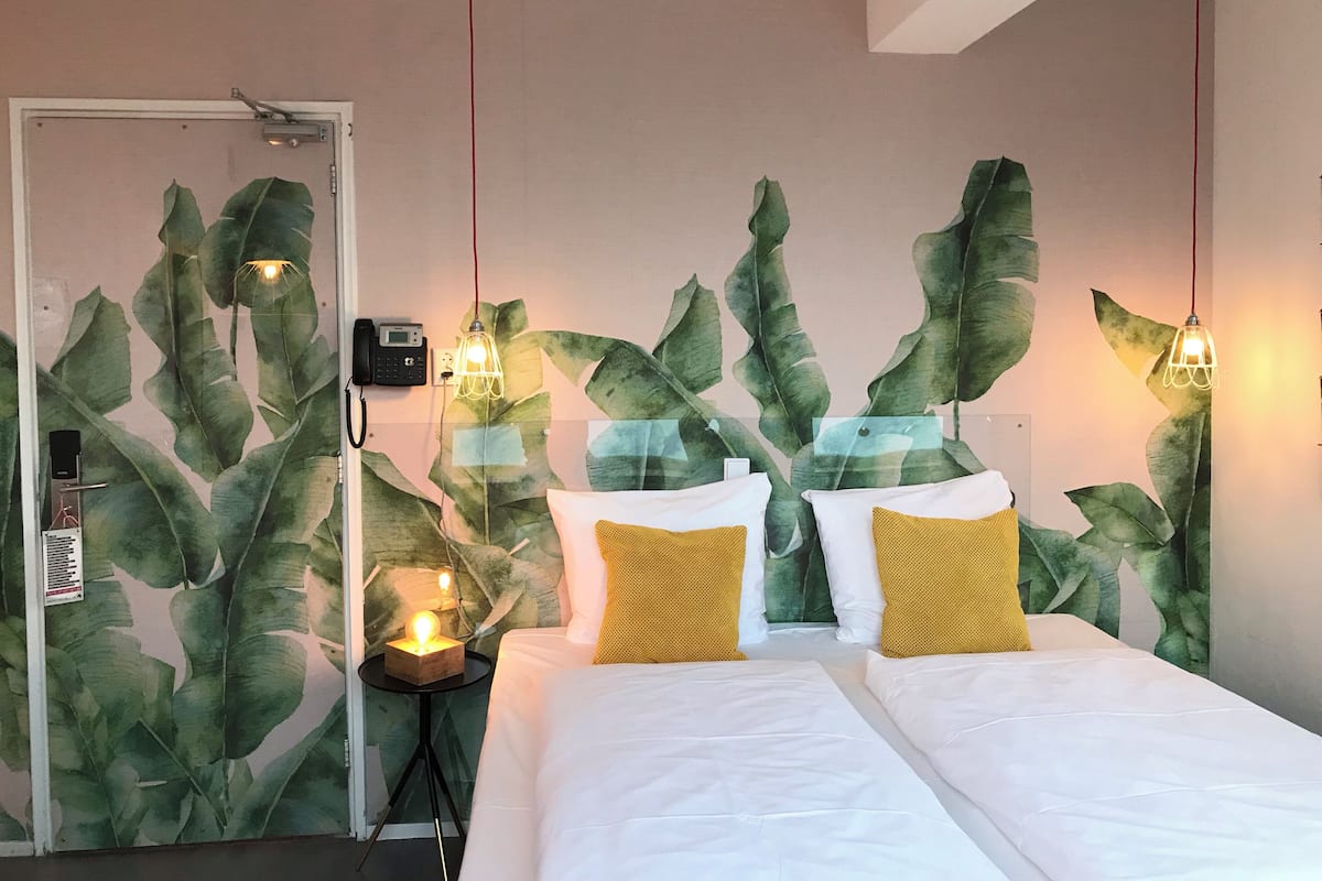 A hotel room with a bed, two yellow pillows, and a wall with large green leaves.