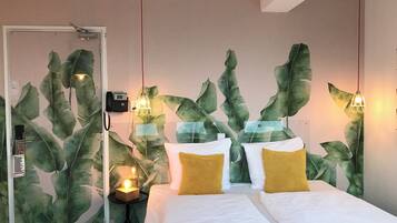 Una habitación de hotel con una cama, dos almohadones amarillos y una pared decorada con hojas verdes grandes.