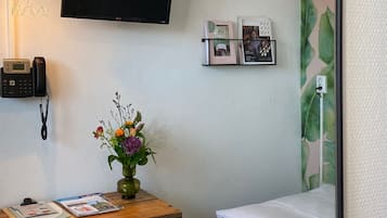 A small room with a desk, chair, bed, TV, and a wall-mounted shelf with photos.