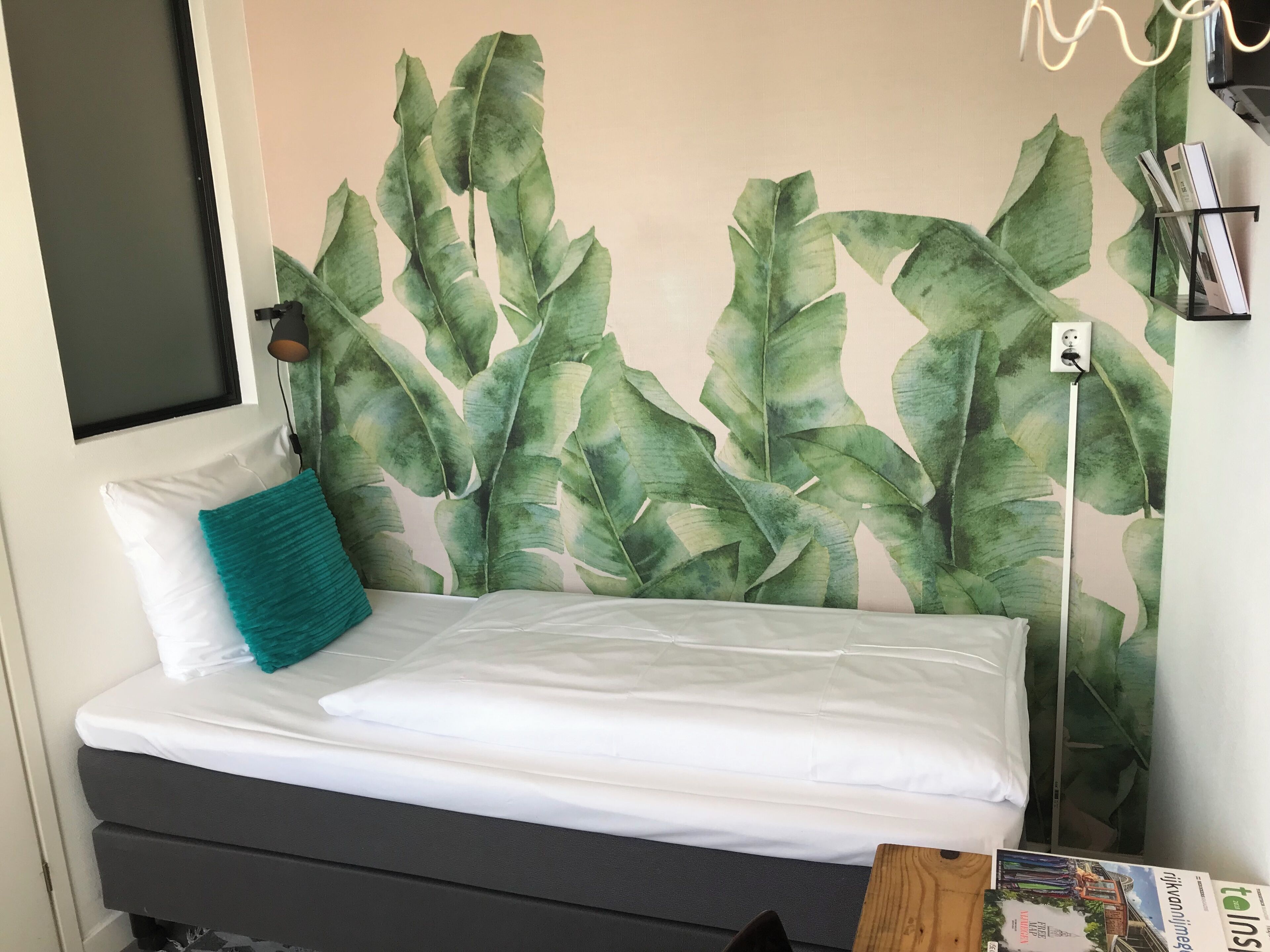 Een bed met wit beddengoed, een blauwgroen kussen en een muur met een groot bladpatroon.