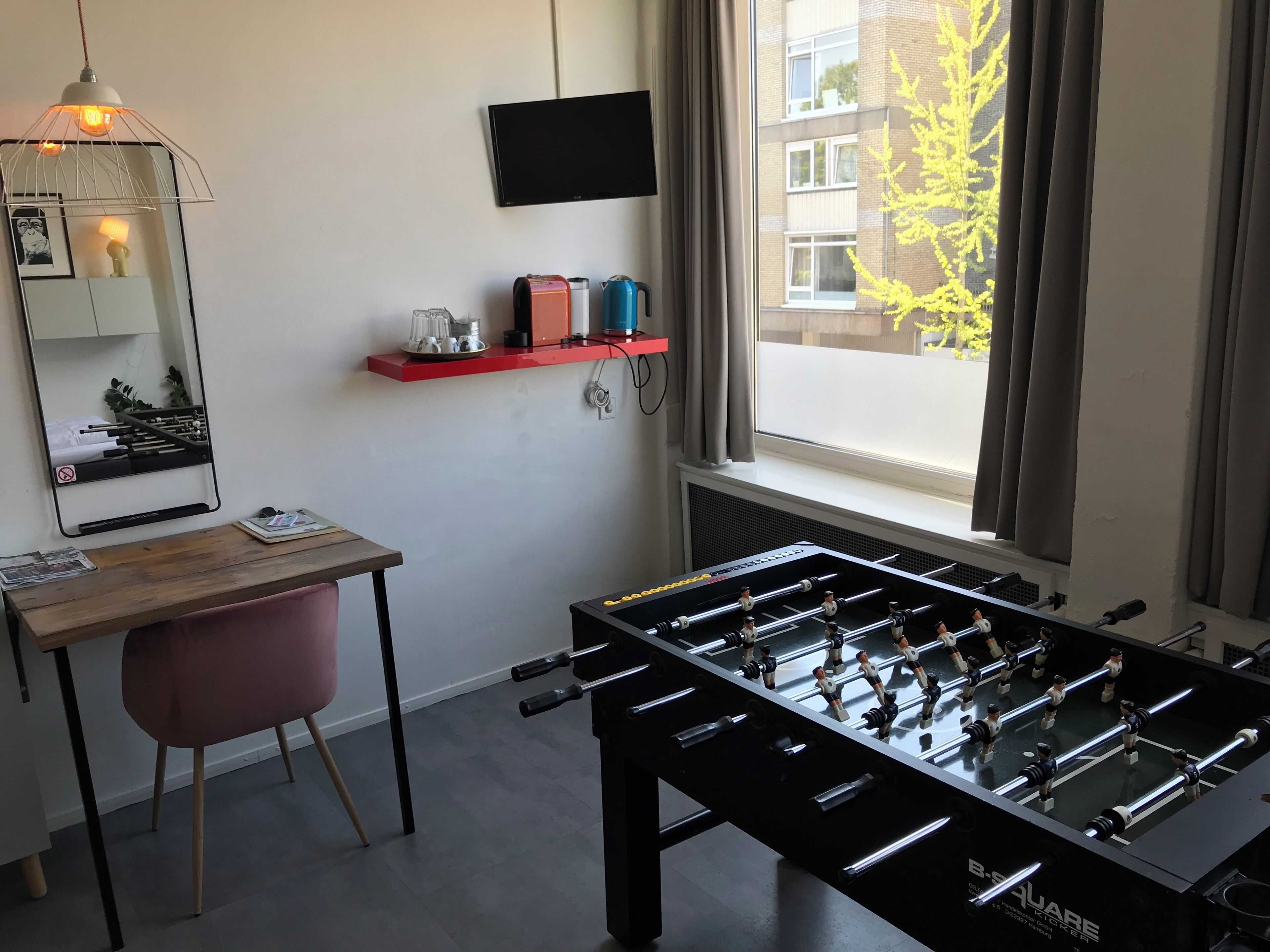 Ein Zimmer mit Kicker, Schreibtisch mit Stuhl, Spiegel, Regal mit Dekoration und Fenster mit Blick auf Gebäude und Bäume.