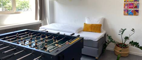 Una habitación de hotel moderna con una mesa de fútbol de mesa, una cama con ropa blanca y una almohada color mostaza, un sofá gris, una planta en maceta y carteles motivacionales en la pared.