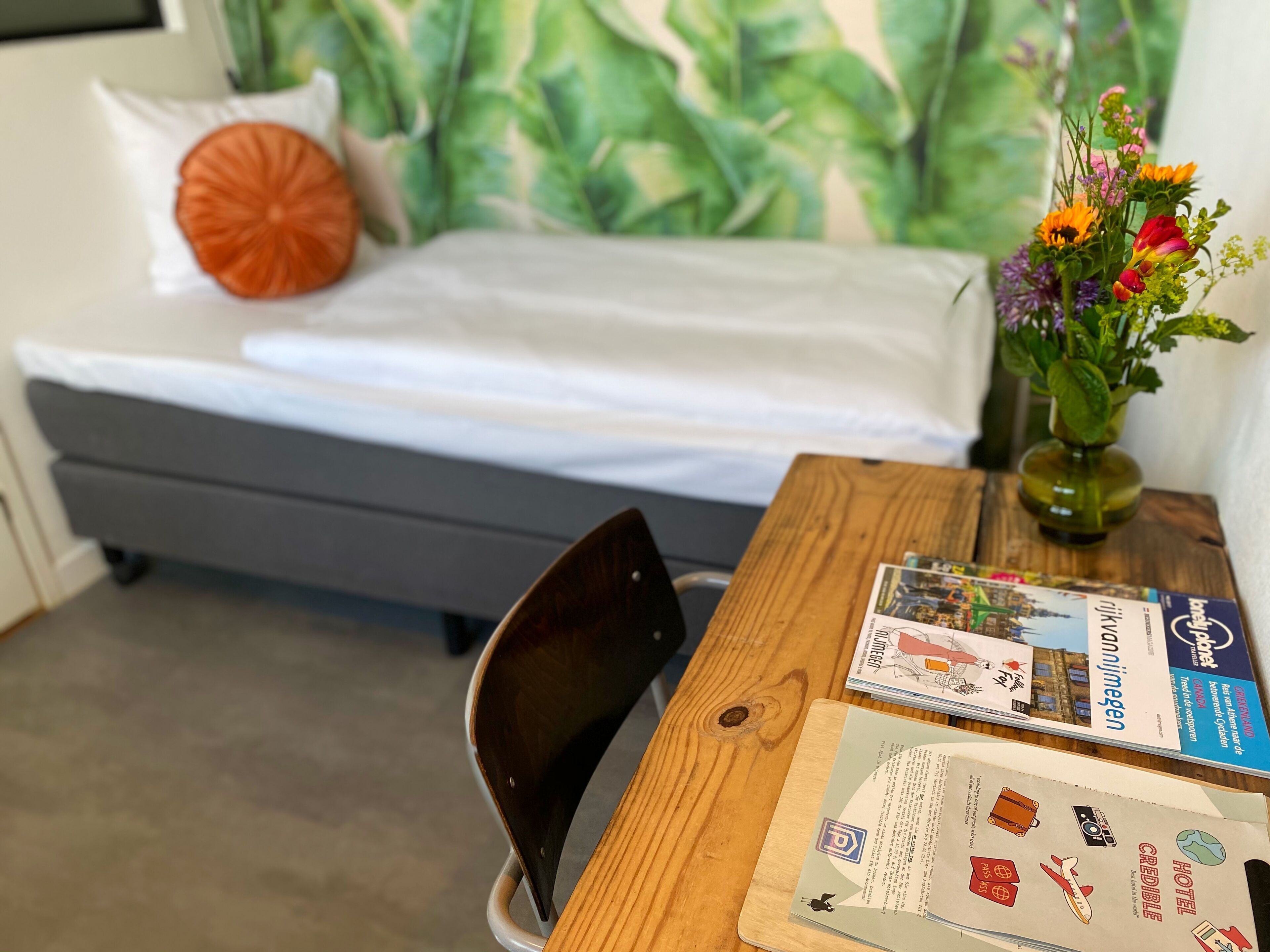 Een slaapkamer met een bed, een houten tafel met brochures, een stoel en een vaas met bloemen.