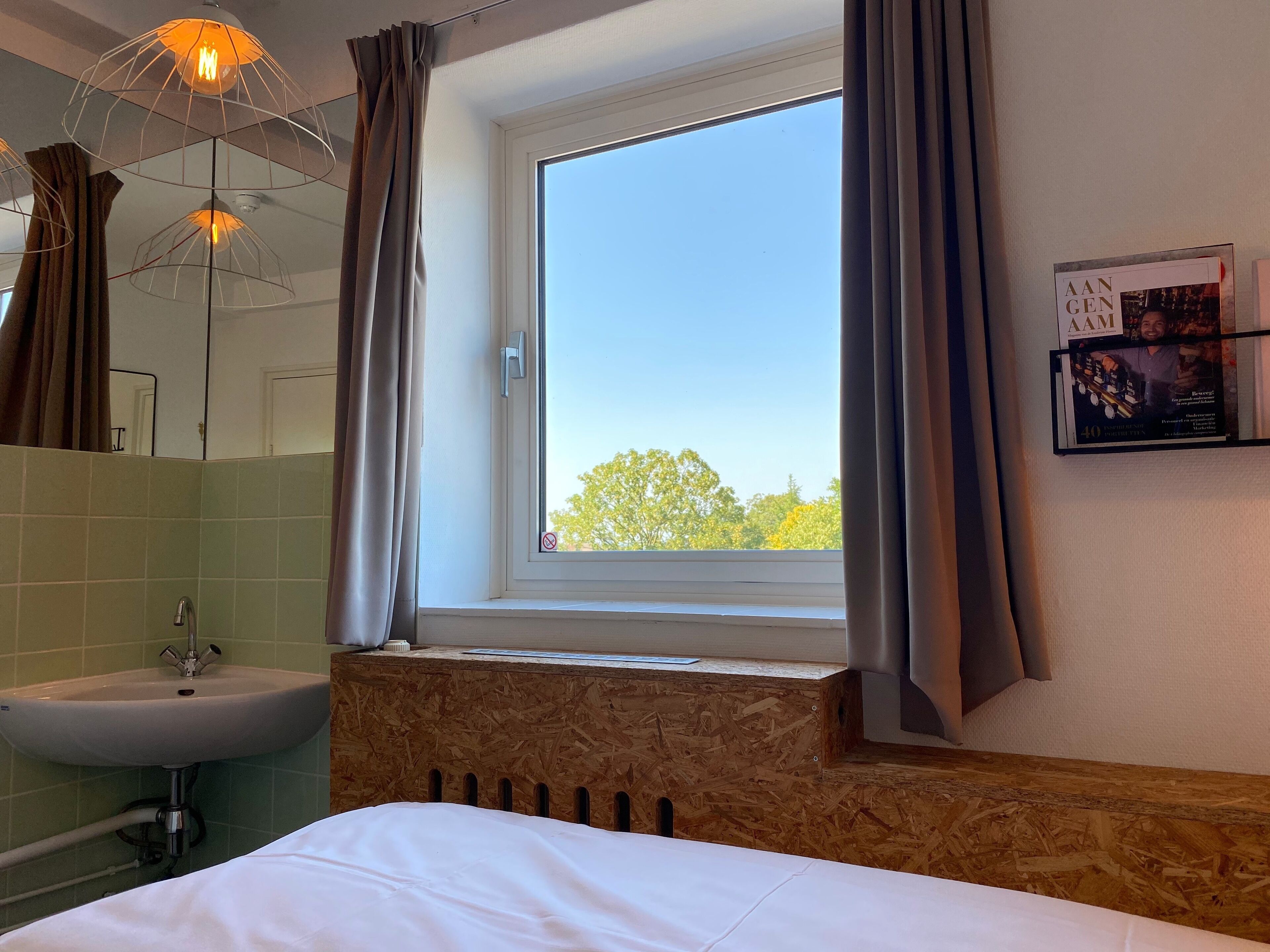 Quarto de hotel com uma janela grande, cabeceira de madeira, cama branca, pia e espelho.