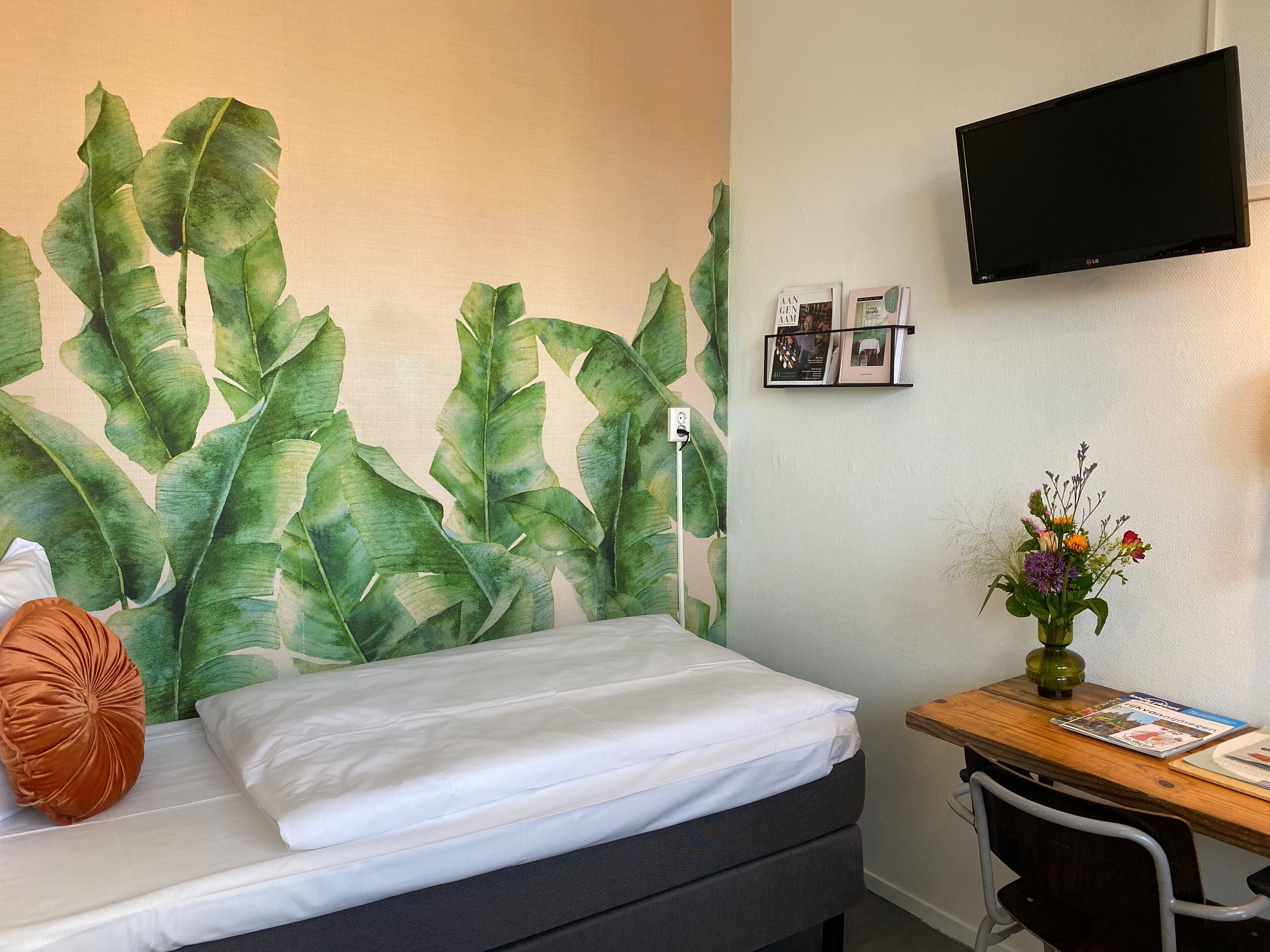 A hotel room with a bed, a desk with a chair, a flat-screen TV mounted on the wall, and a large wall mural of green leaves.