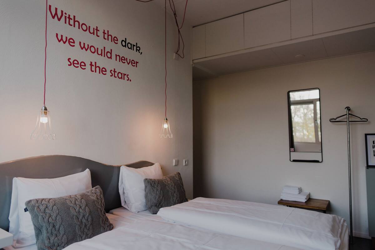 A modern hotel room with a large bed, a bedside table, a wall-mounted mirror, and a motivational quote on the wall.