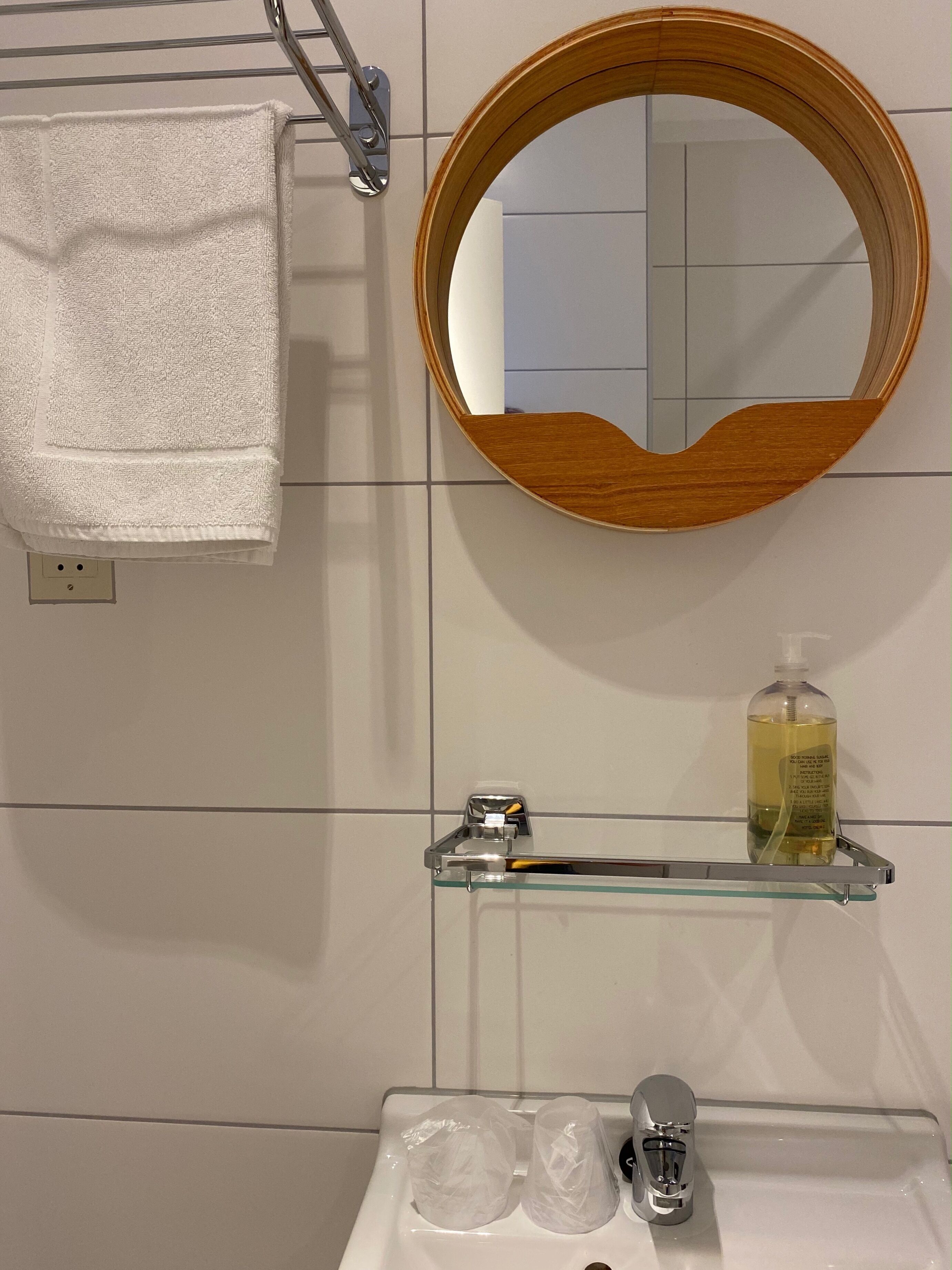 A bathroom with a white towel hanging on a rack, a wooden-framed mirror, a glass shelf with a bottle of liquid soap, and a sink with a faucet.
