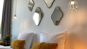 Ein ordentlich bezogenes Bett mit weißen Leinen und zwei senfgelben Kissen, eine Wand mit drei Spiegeln und einer hängenden Leuchte.