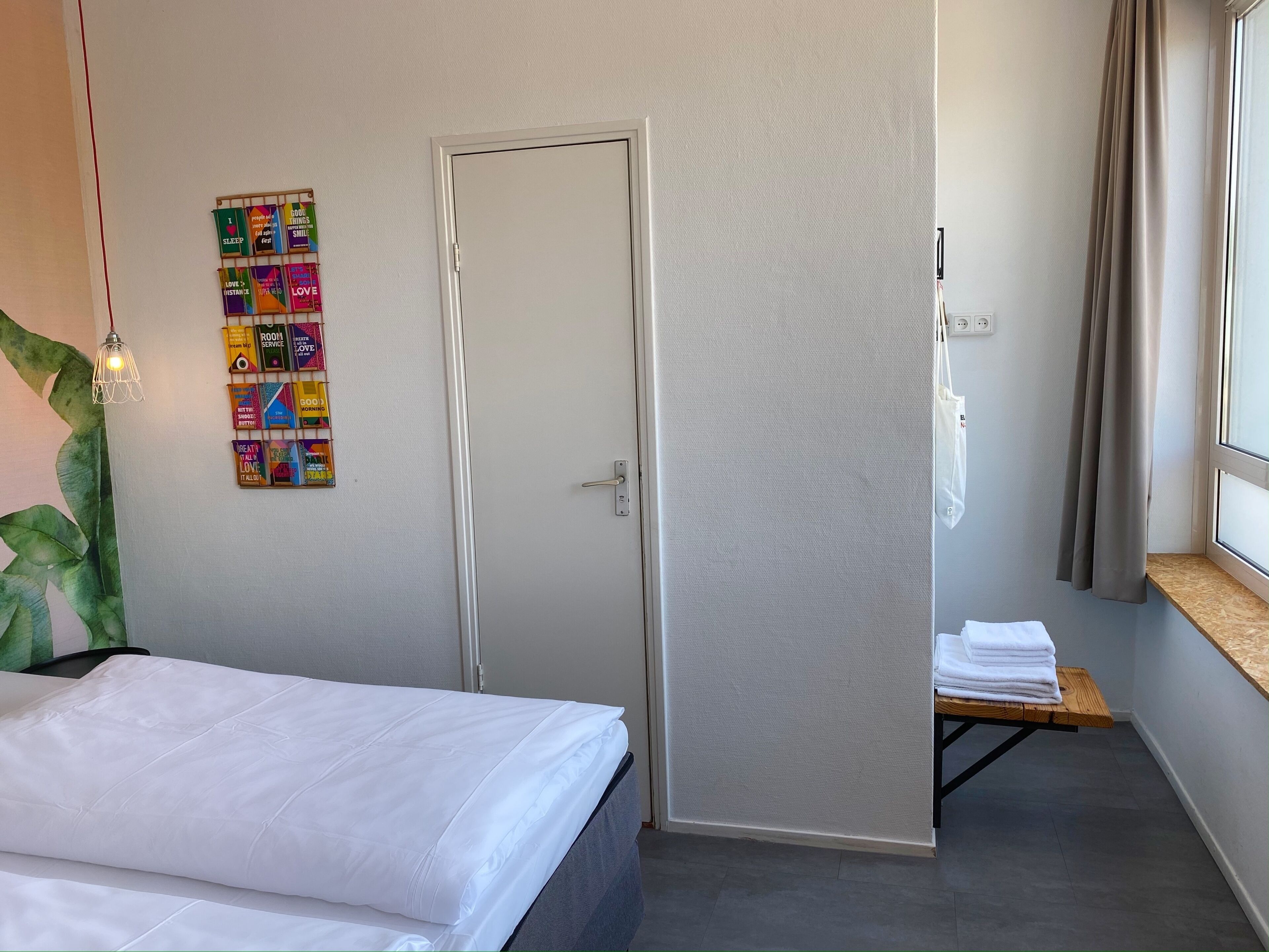 A hotel room with a bed, a door, a small table with towels, a wall-mounted light fixture, and a window with curtains.