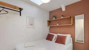 Una cama bien hecha con sábanas blancas y dos almohadas marrones, un estante de madera con plantas, un cuadro en la pared y un espejo que refleja la puerta.